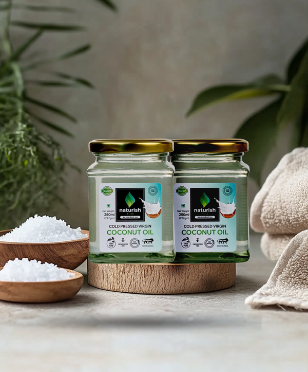 Two Naturish Elite cold pressed virgin coconut oil glass jars arranged on a wooden stand with bath salts, towels and green leaves in a spa setting.
