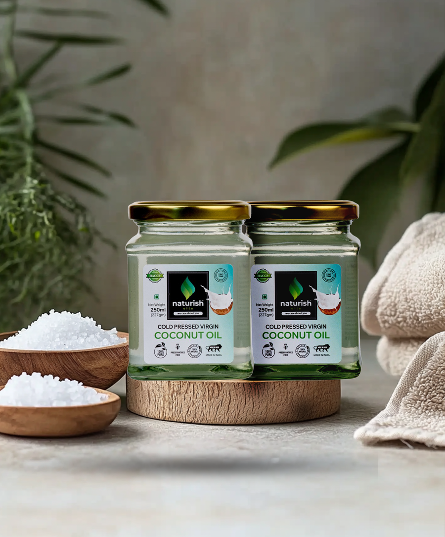 Two Naturish Elite cold pressed virgin coconut oil glass jars arranged on a wooden stand with bath salts, towels and green leaves in a spa setting.