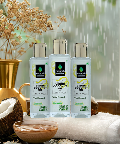 Three Naturish Elite cold pressed virgin coconut oil bottles for skin and hair care arranged on a spa table with towels, coconut shells and a bowl of oil against a rainy window.