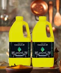 Two bright yellow 2‑litre Naturish coconut oil cans with black labels on a rustic kitchen table, copper utensils blurred in background.