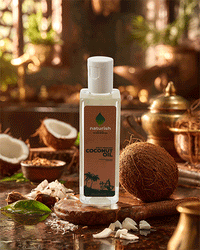 Small Naturish cold-pressed coconut oil bottle with flip cap surrounded by coconuts and brassware on a rustic kitchen counter.